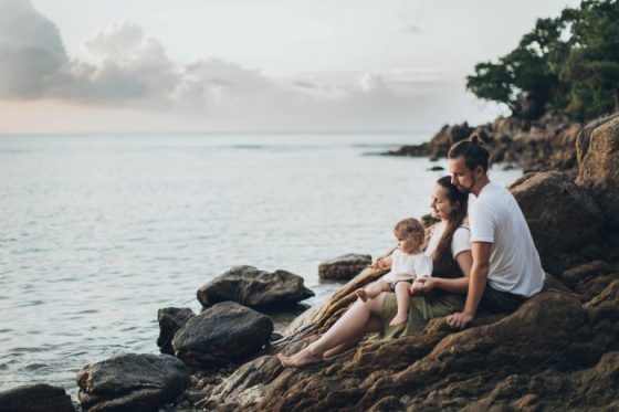A family of three sitting on rocks by the ocean.