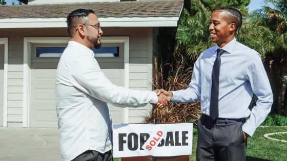 Two men shaking hands with a for sale signed stating sold between them.